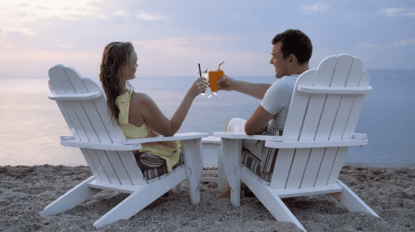 Couple Sitting On Adirondack Chairs Having A Toast In Front Of The Sea
