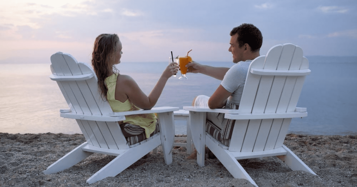 Couple Sitting On Adirondack Chairs Having A Toast In Front Of The Sea