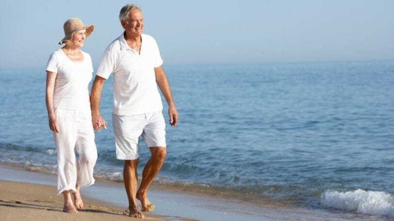 Elderly Couple Dressed In White Holding Hands And Walking By The Sea Shore