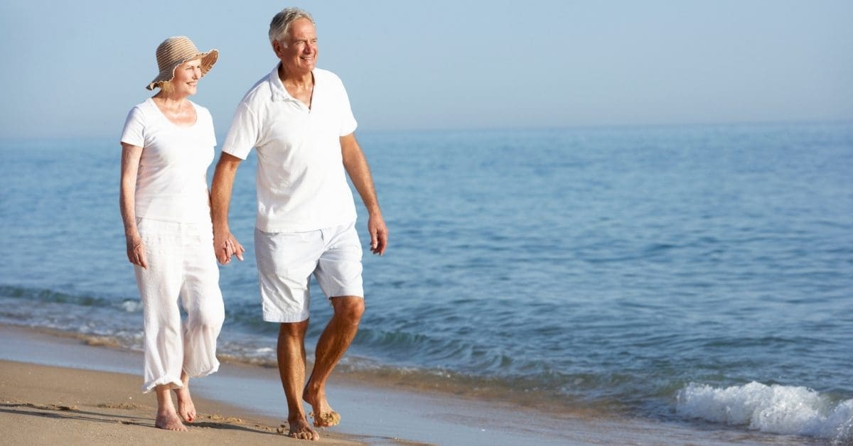 Elderly Couple Dressed In White Holding Hands And Walking By The Sea Shore